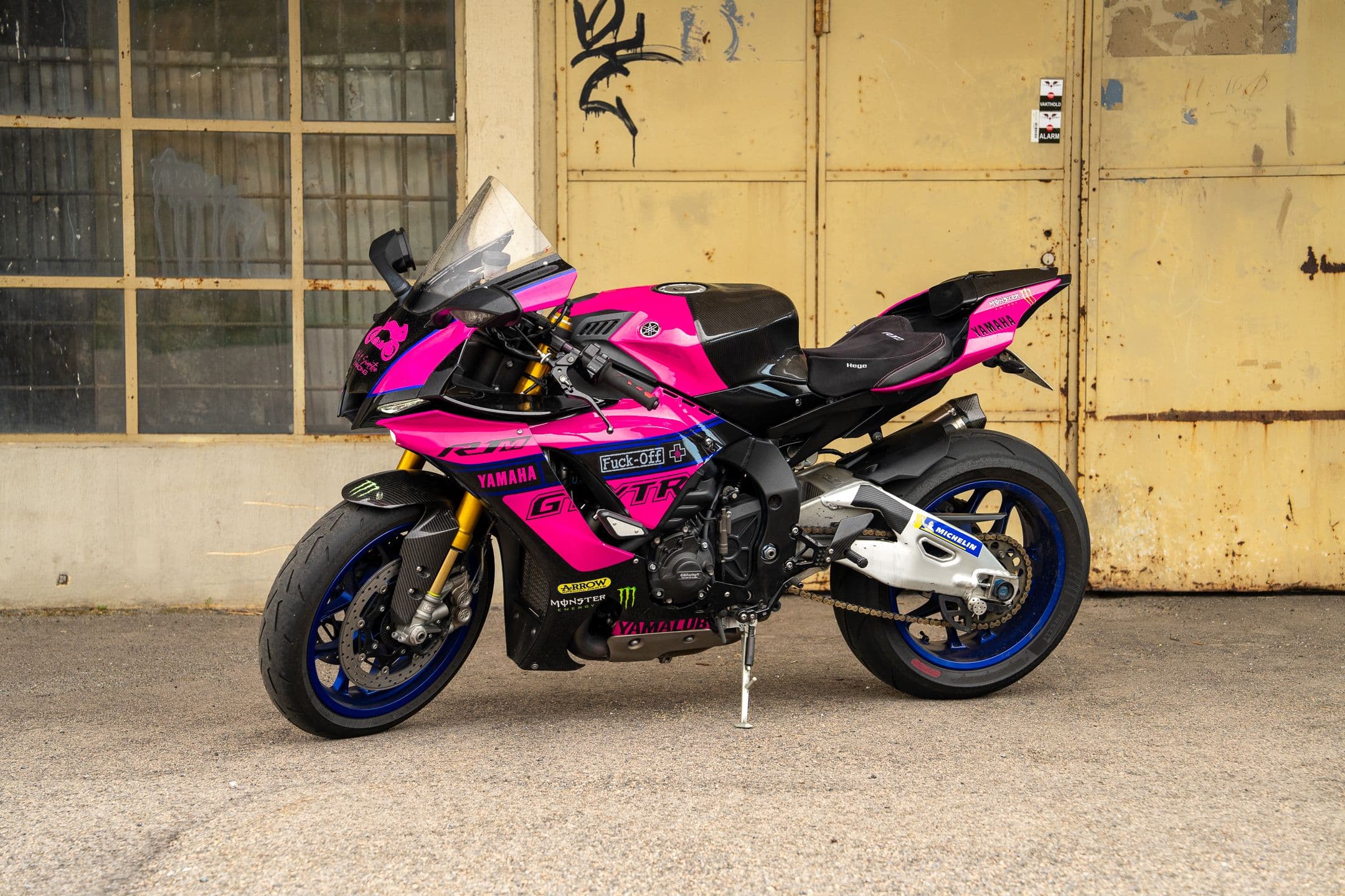 Rosa og svart Yamaha R1M sett skrått forfra, parkert på sidestøtten foran en gammel, gul garasjeport
